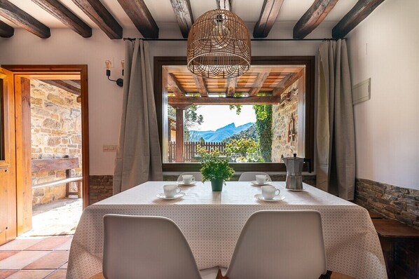 A dining area with a table set for four, a hanging light fixture, and a view of a stone wall and mountains. - Fidalsa House of Views (Cabezon de Liebana)
