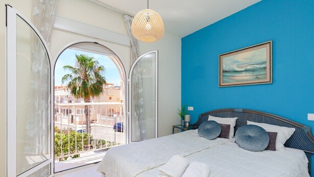 A bedroom with a bed, a painting, and a view of a palm tree and buildings through a glass door. - Goldencore by Fidalsa (Orihuela)