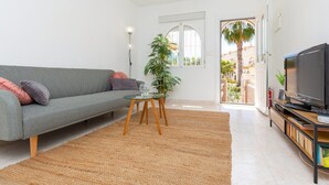 A modern living room with a grey sofa, a small round table, a television on a wooden stand, and a large window with a view of palm trees and buildings. - Goldencore by Fidalsa (Orihuela)