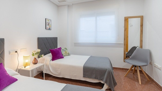 Habitación de hotel con dos camas, una mesita de noche con lámpara, una silla, una ventana y un espejo.