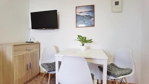 Ein Zimmer mit einem an der Wand befestigten Fernseher, einem gerahmten Bild, einem kleinen Tisch mit einer Pflanze und einem Schrank.