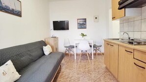 Ein kompakter Wohnbereich mit einer Couch, einem kleinen Esstisch mit Stühlen, einem an der Wand montierten Fernseher und einer Küchenzeile mit Schränken und Spüle.