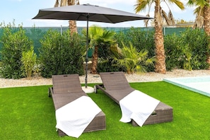 Two reclining lounge chairs with white covers, a large umbrella, and a pool in the background. - Fidalsa Divine Hiding (Algorfa)