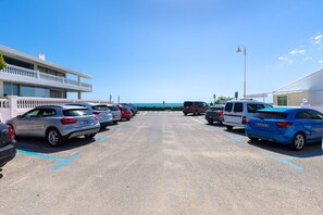 A parking lot with multiple cars, a building with a balcony, and a clear view of the sea in the background.