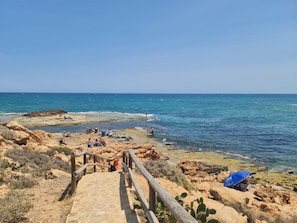 A coastal beach with a wooden walkway leading to the water, rocky terrain, and people enjoying the beach. - Fidalsa Moon Dunes Villa (Torrevieja)