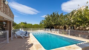 Une piscine rectangulaire dont les bords sont recouverts de carreaux, entourée d’une clôture blanche et située à côté d’un bâtiment à la façade en briques.