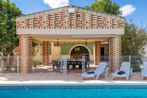 Ein Poolbereich mit überdachter Sitzgruppe, ein Pool mit dekorativem Rand und ein Backsteinhaus mit der Jahreszahl '1922' an der Fassade.