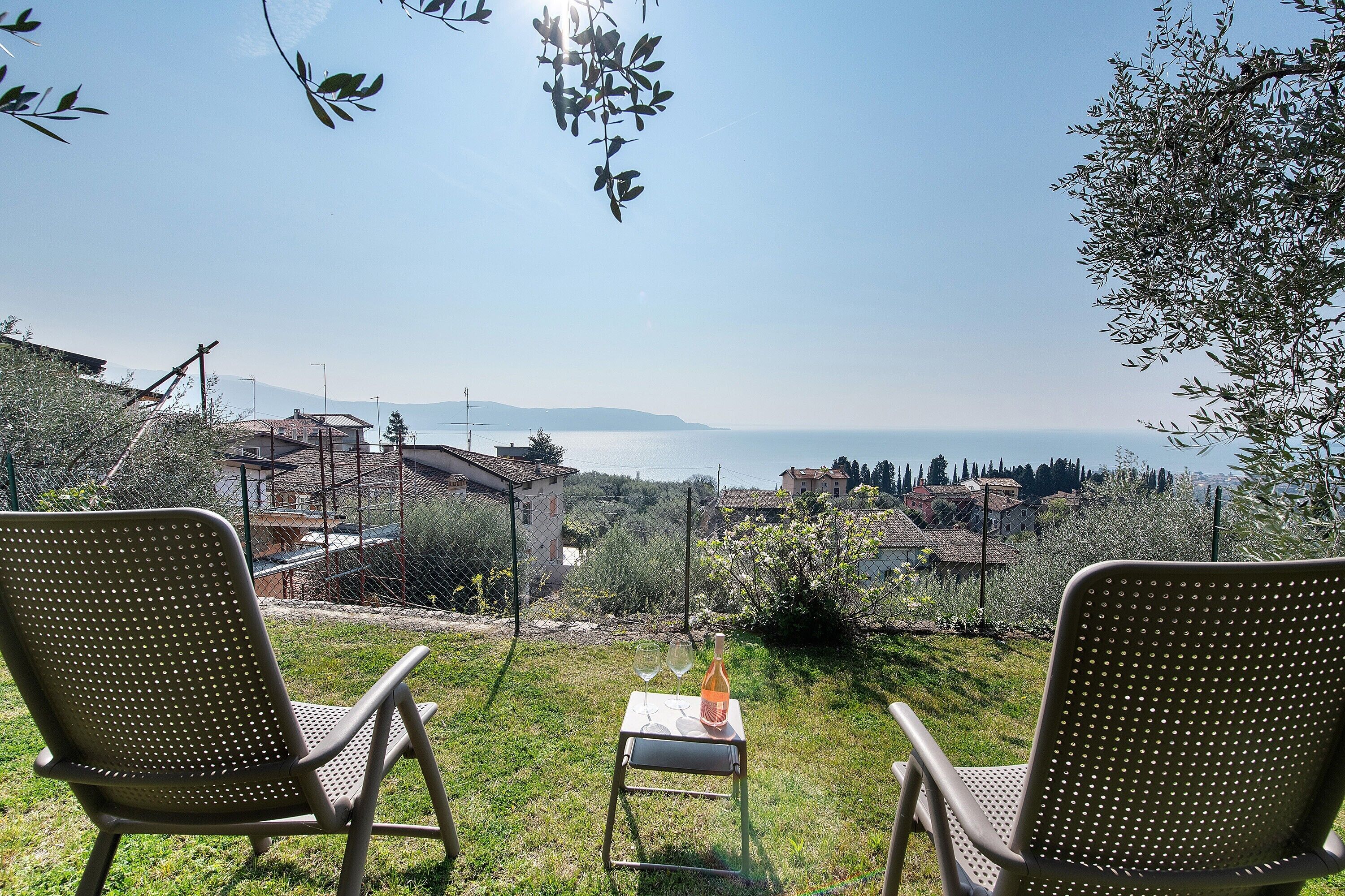 Deux chaises sont placées face à une table sur laquelle se trouvent une bouteille et des verres. L’ensemble offre une vue panoramique sur une zone côtière, avec des maisons et de la végétation.