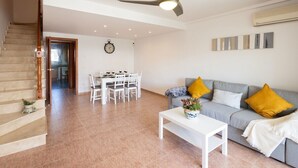 A spacious living area with a staircase, a dining table with chairs, a gray sofa with yellow cushions, a white coffee table, and a ceiling fan. - Golden Coast Retreat by Fidalsa (Creixell)