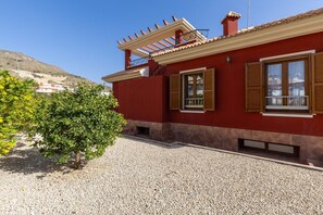 Un bâtiment rouge avec une allée gravillonnée, des volets en bois et une pergola.
