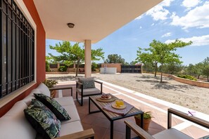 Un espacio de estar al aire libre cubierto, con un sofá, sillas y una mesa con una bebida y fruta.