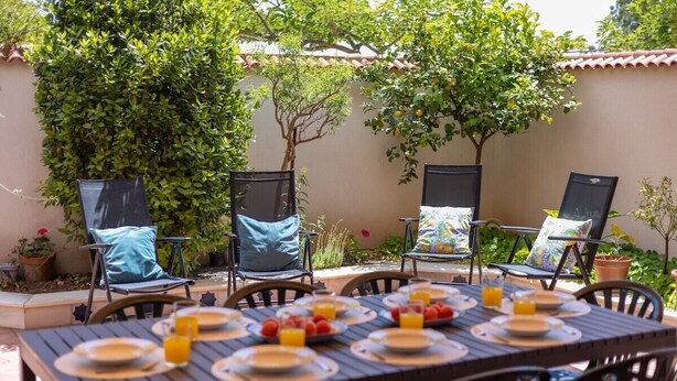 Une terrasse extérieure avec une table dressée pour le petit-déjeuner, entourée de chaises dotées de coussins.