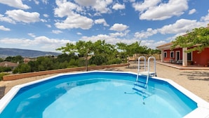 Une piscine rectangulaire à bordure blanche, entourée d’une zone sablonneuse, avec un ciel bleu clair et quelques nuages épars au-dessus.