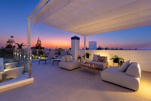 A modern outdoor living space with a glass balustrade, a white sofa set, a wooden coffee table, and a view of the city skyline at dusk. - Dreamscape Villa by Fidalsa (Los Alcazares)