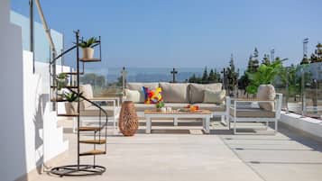 A rooftop terrace with a sofa, coffee table, and chairs, surrounded by potted plants and a decorative vase.
