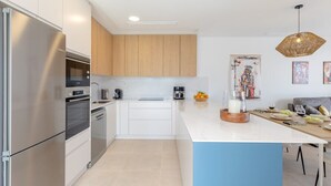 A modern kitchen with stainless steel appliances, wooden cabinets, a marble countertop, and a dining area with a table set for two. - Dreamscape Villa by Fidalsa (Los Alcazares)