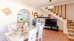 Un comedor con una mesa blanca dispuesta para cuatro personas, un televisor y una escalera que sube a una plataforma.