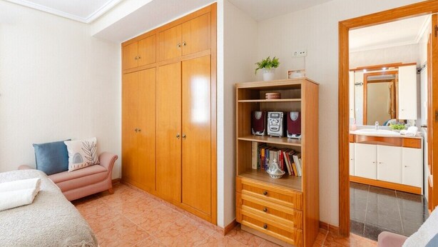 Un dormitorio con cama, armario, estante con libros y una planta, y un baño con espejo y lavamanos.