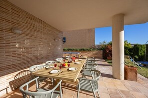 Un espace extérieur aménagé pour les repas, avec un mur en briques, une longue table en bois et des chaises blanches.