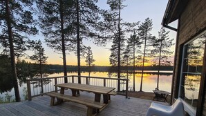 Eine Holzterrasse mit Picknicktisch und Stühlen, mit Blick auf einen stillen See, Bäume und einen Sonnenuntergang.