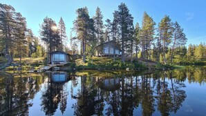 Eine Holzhütte am Seeufer mit Spiegelbild im Wasser, umgeben von hohen Bäumen und blauem Himmel.