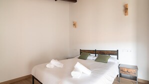 Una cama bien hecha con ropa blanca y toallas enrolladas, cabecera de madera, una mesita de noche pequeña y un aplique de pared.