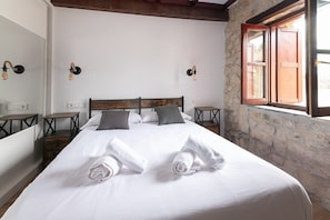 A neatly made bed with towels, a stone wall, a wooden ceiling, and a window with wooden shutters. - Infinity Coast II by Fidalsa (Miengo)