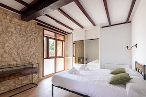 A bedroom with a stone wall, wooden beams, a large bed with pillows, a bedside table, and a mirror. - Infinity Coast I by Fidalsa (Miengo)