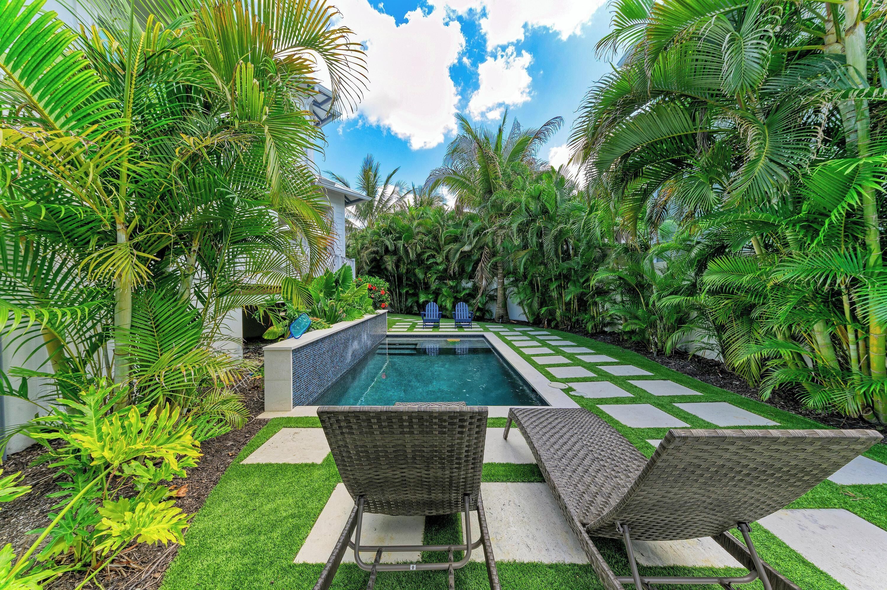 A swimming pool with two wicker lounge chairs, surrounded by lush greenery and a clear sky.