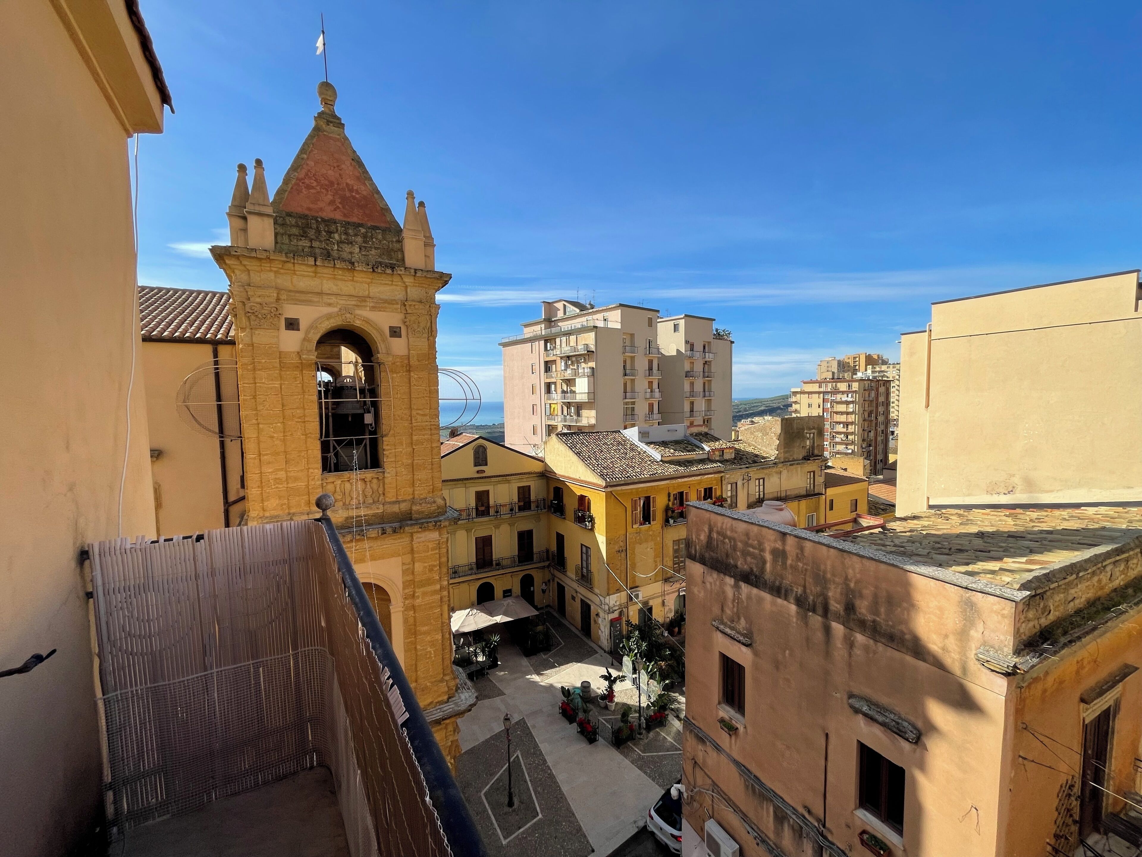 A courtyard with a bell tower, surrounded by buildings with balconies and a clear sky.