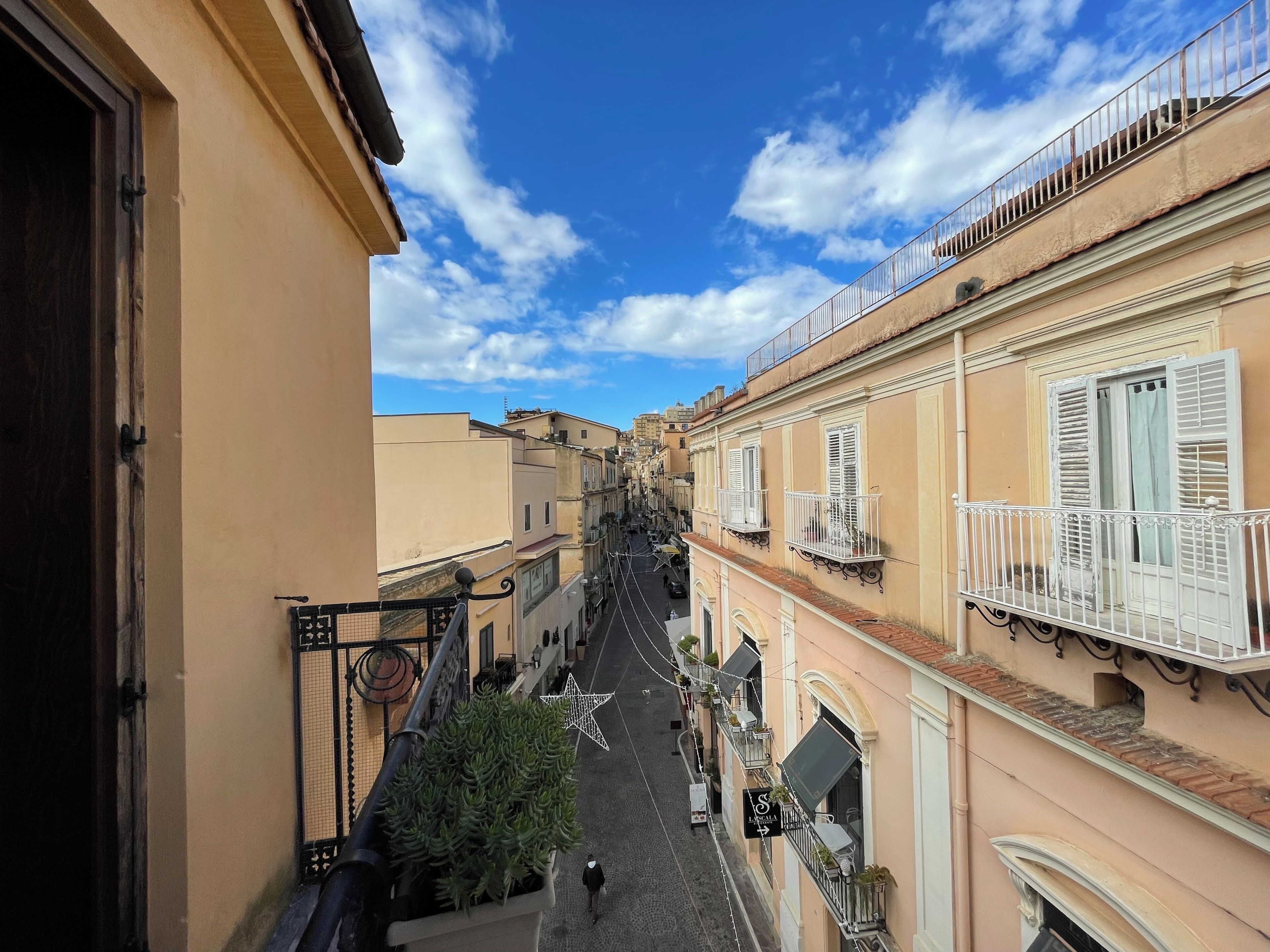 A narrow street with buildings on either side, featuring balconies with railings and potted plants.