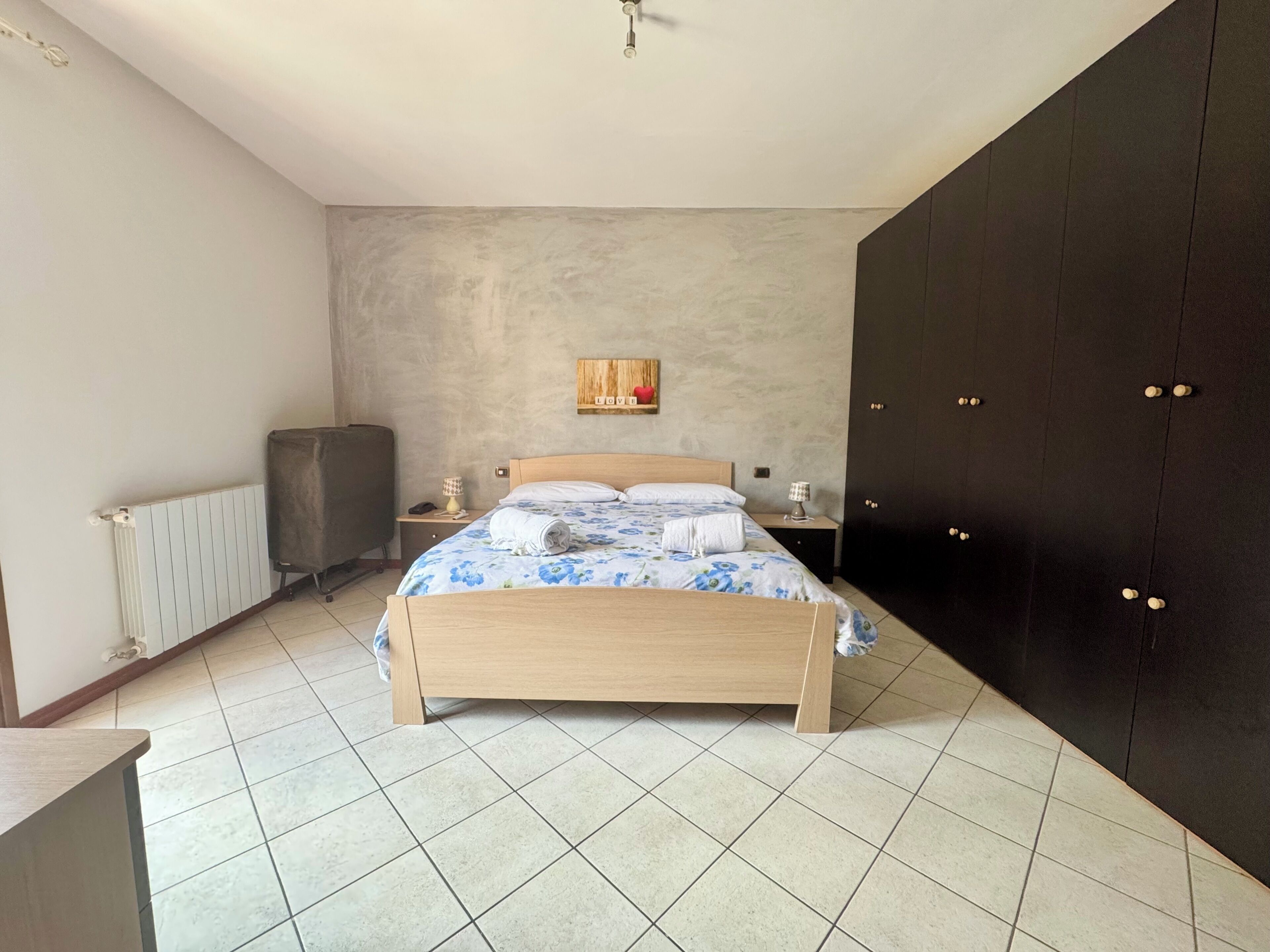 Une chambre comprenant un lit, un radiateur, un bureau, une chaise, une armoire et une étagère fixée au mur, sur laquelle est posé un panneau.