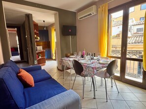 A living room with a sofa, dining table set for four, a TV, and a balcony with yellow curtains. - Akragas home 2 with city view (Agrigento)