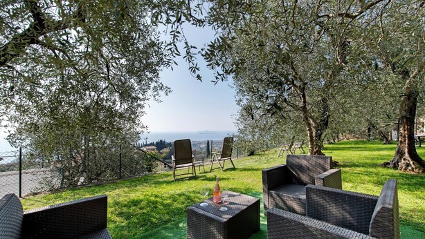 An outdoor seating area with wicker furniture, a bottle, and a glass, surrounded by trees and a view of the sea.