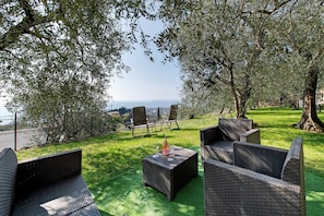 Un espace extérieur aménagé avec des meubles en osier, une bouteille et un verre, entouré d’arbres et offrant une vue sur la mer.