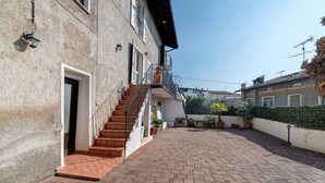 A building with a staircase leading to a door, a paved courtyard, and a hedge.