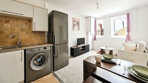 A compact kitchen with a washing machine, a refrigerator, and a dining area with a table set for two. - Chic 1BR West Ealing - 15m Heathrow - 20m City (London)