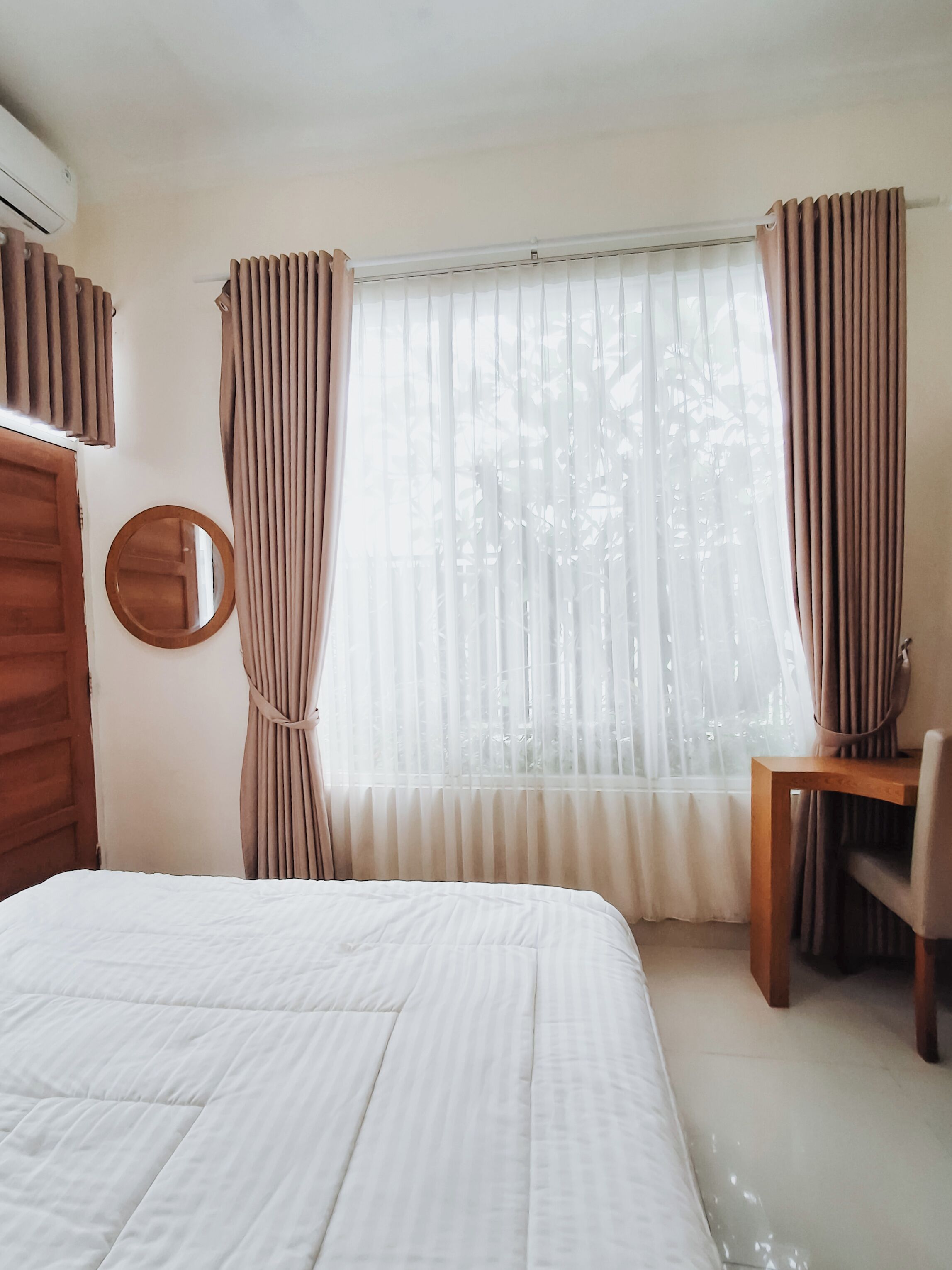 A bedroom with a bed, a round mirror, a wooden desk, and a window with curtains.