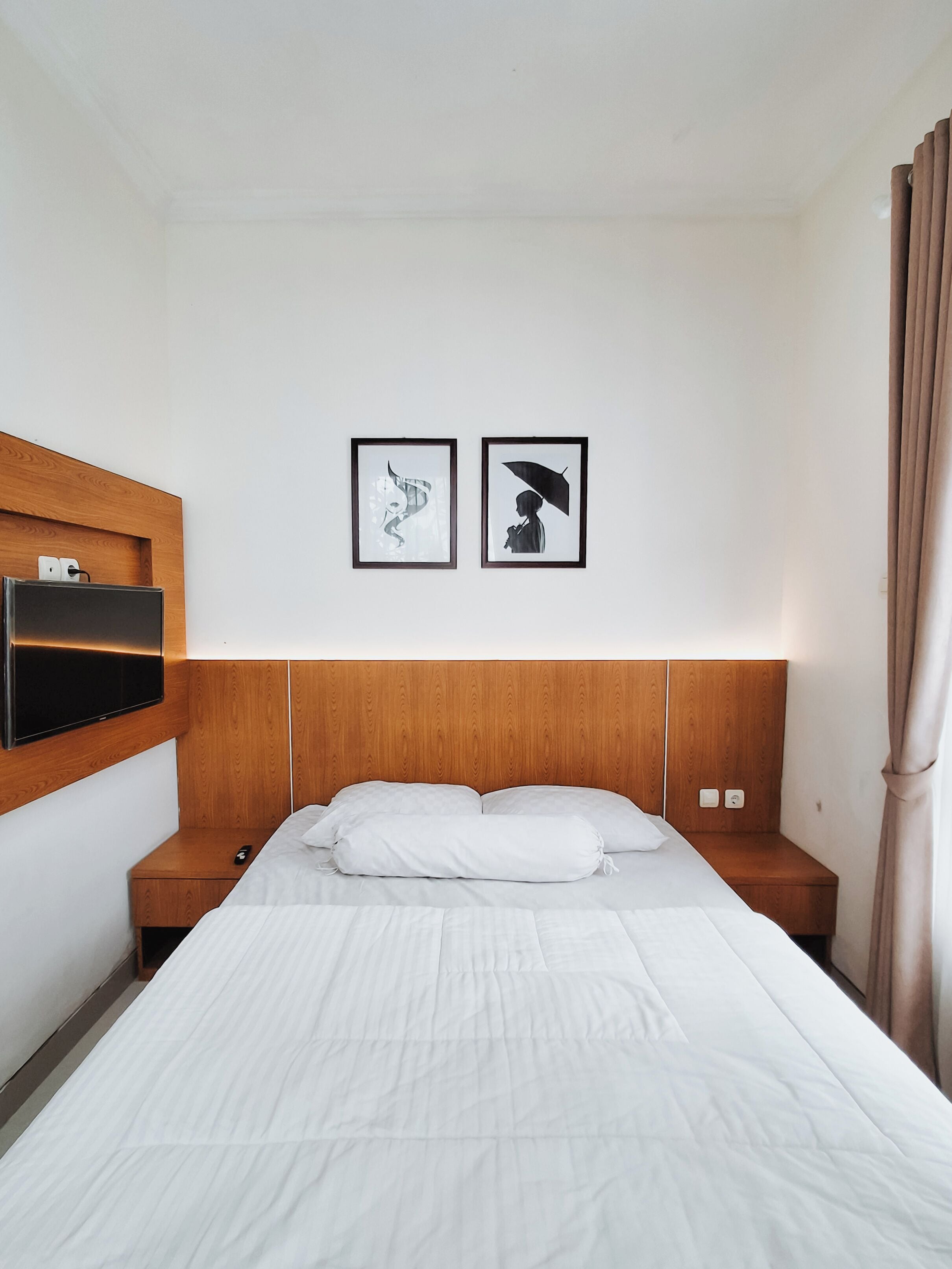 A hotel room with a wooden headboard, a bed with white bedding, a mounted TV, and two framed pictures on the wall.