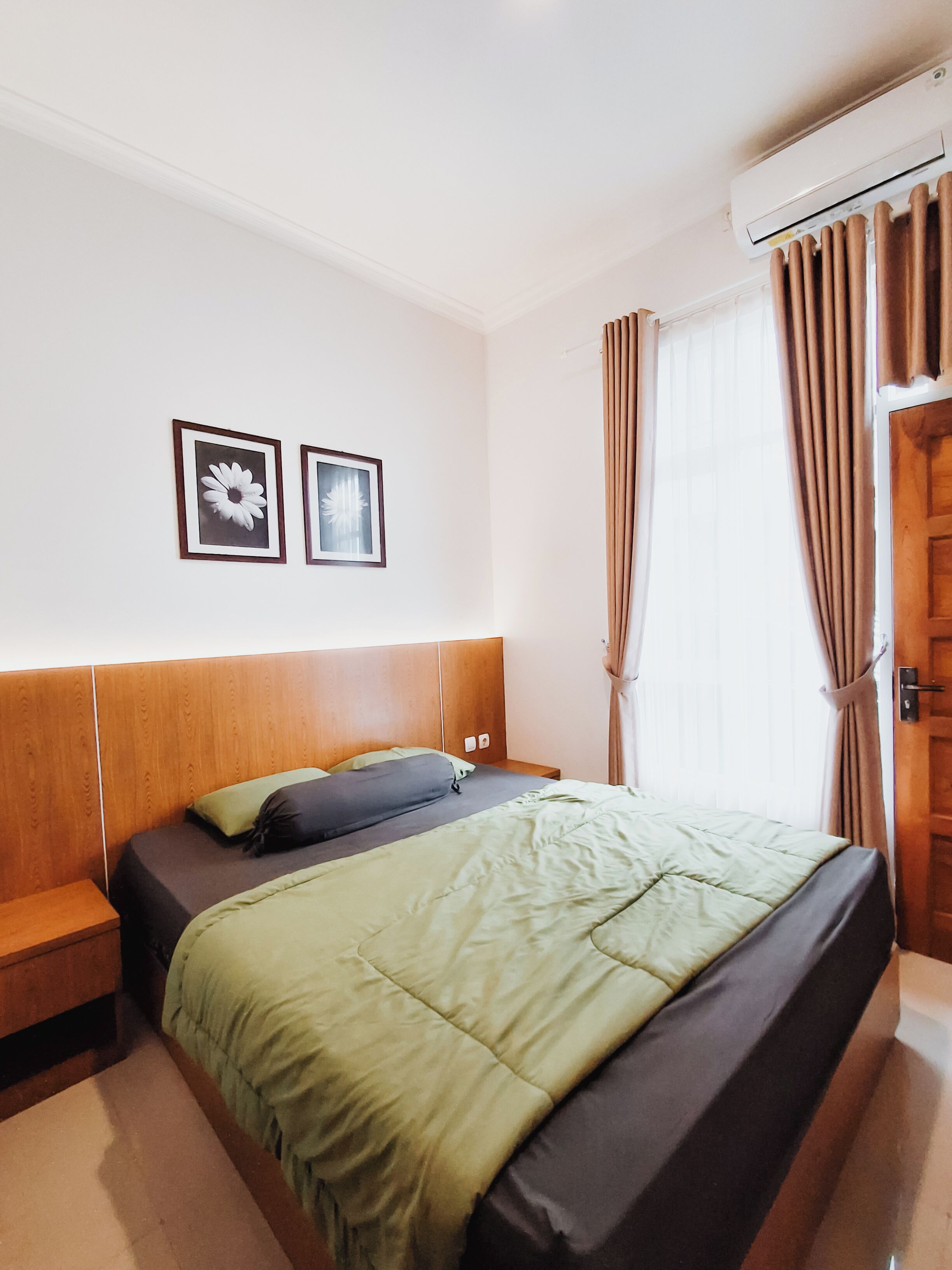 A hotel room with a wooden headboard, a bed with green and black bedding, two framed pictures on the wall, a window with curtains, and an air conditioning unit.
