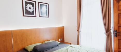 A hotel room with a wooden headboard, a bed with green and black bedding, two framed pictures on the wall, a window with curtains, and an air conditioning unit.