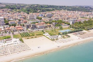 Vista aerea di una cittadina costiera con spiaggia, edifici di un hotel e un ponte sullo sfondo.