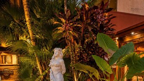Una estatua en un jardín con un sendero de ladrillo, rodeada de plantas tropicales y un edificio al fondo.