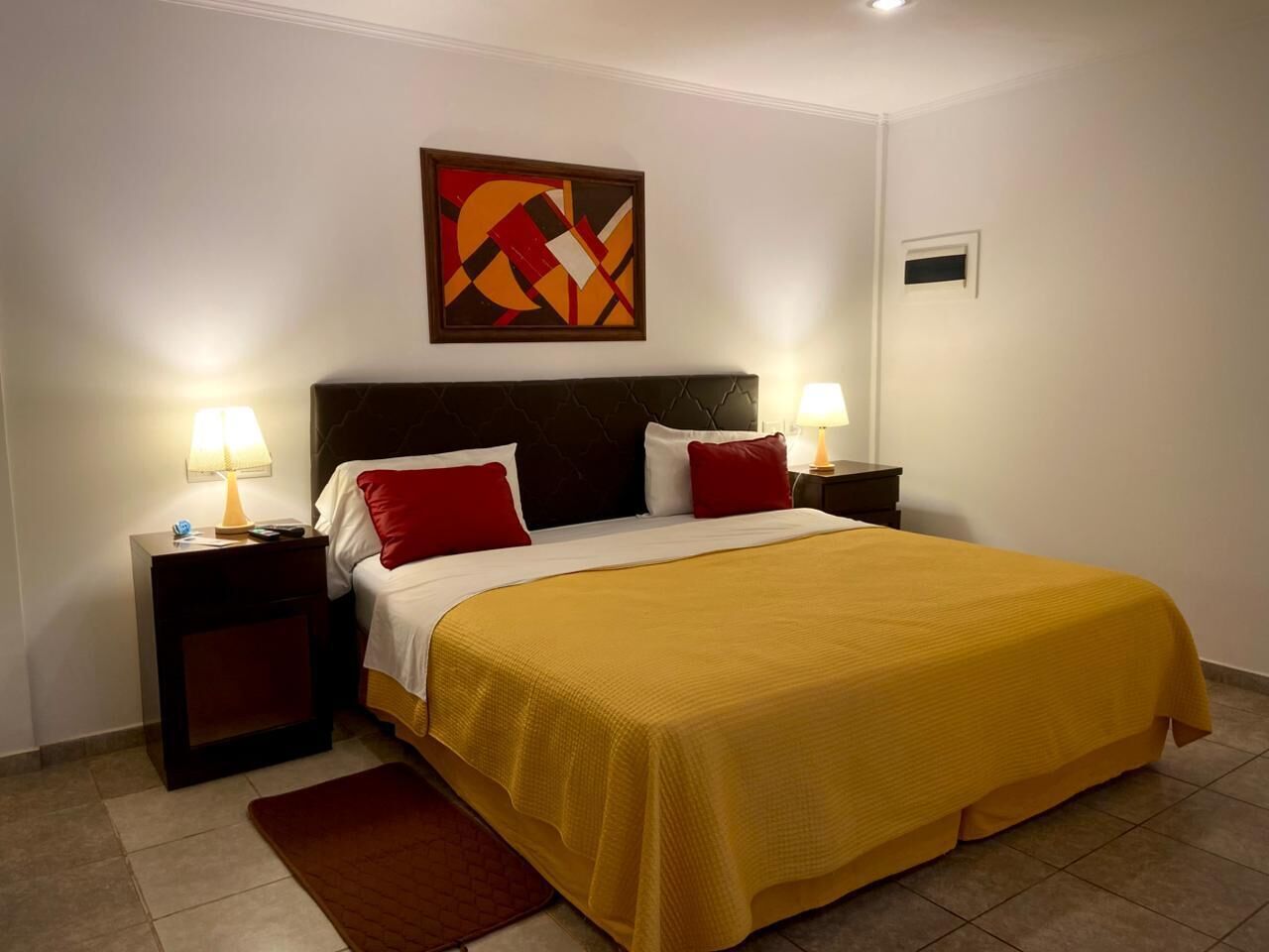 Hotelkamer met een bed, twee nachtkastjes met lampen, een rood-wit kussen, een gele sprei, een rood tapijt en een geometrisch schilderij aan de muur.