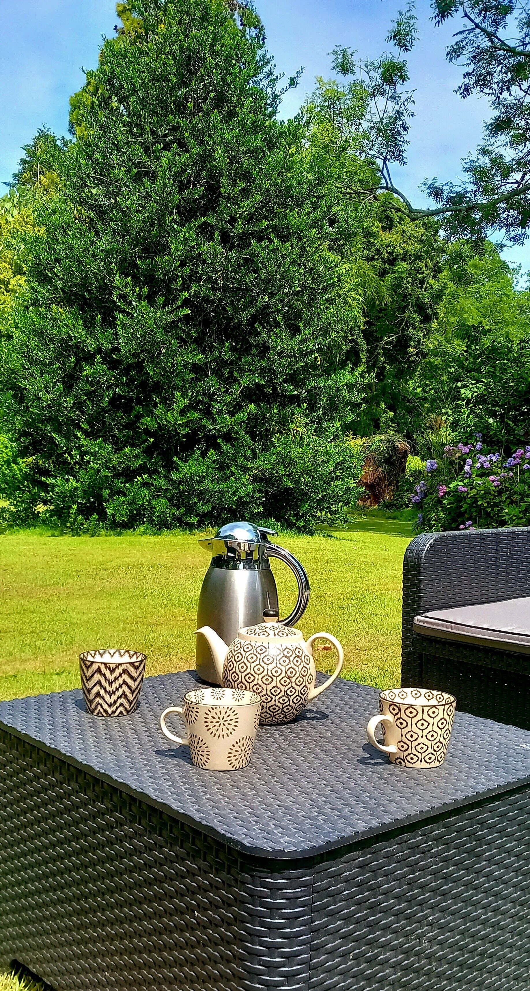 Ein Tisch mit Wasserkocher, Teekanne und verzierten Tassen, draußen auf einer Rasenfläche vor Bäumen.