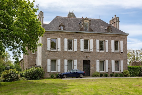 A stone building with multiple windows and shutters, a car parked in front, and a well-maintained lawn.