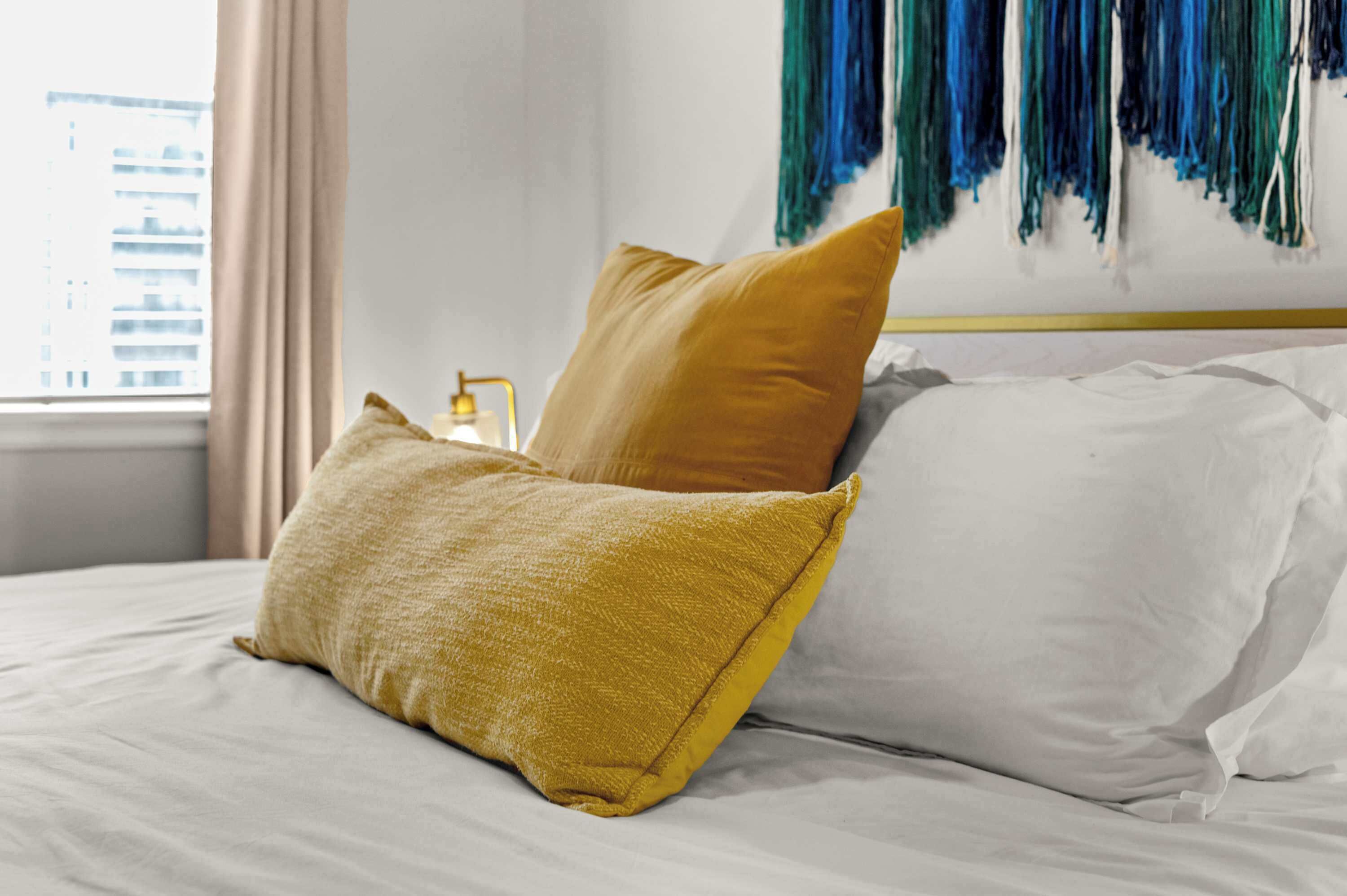 A bed with white and mustard pillows, a bedside table with a lamp, and a wall with blue and green hanging decorations.