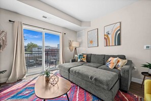 A modern living room with a grey sectional sofa, a round coffee table, a colorful rug, and a balcony with a view of the city.