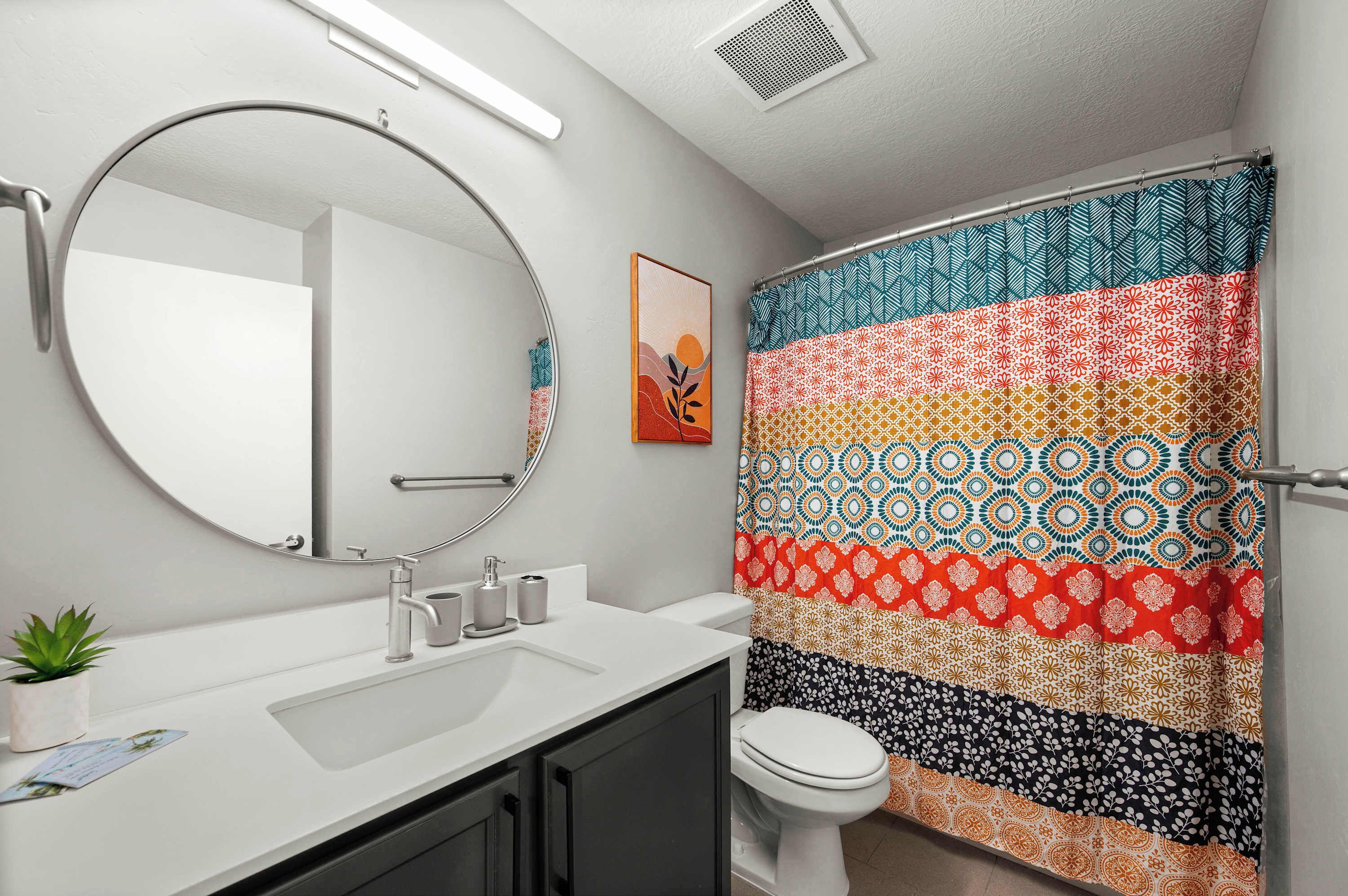 Une salle de bain avec un miroir rond, un lavabo, des toilettes et un rideau de douche coloré.