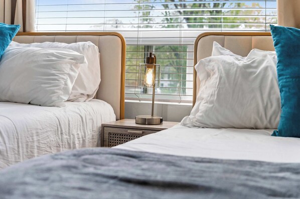 Quarto de hotel com duas camas, uma mesa de cabeceira com abajur e uma janela com persianas.